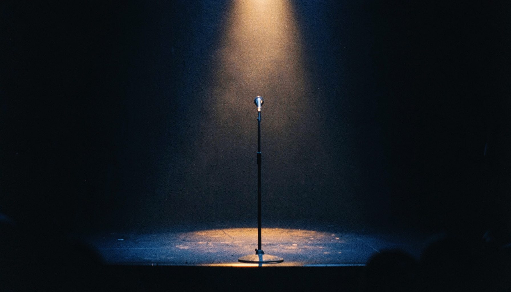 A dimly lit stage with a single microphone stand under a warm spotlight, deep blue and amber tones, theatrical and mysterious mood