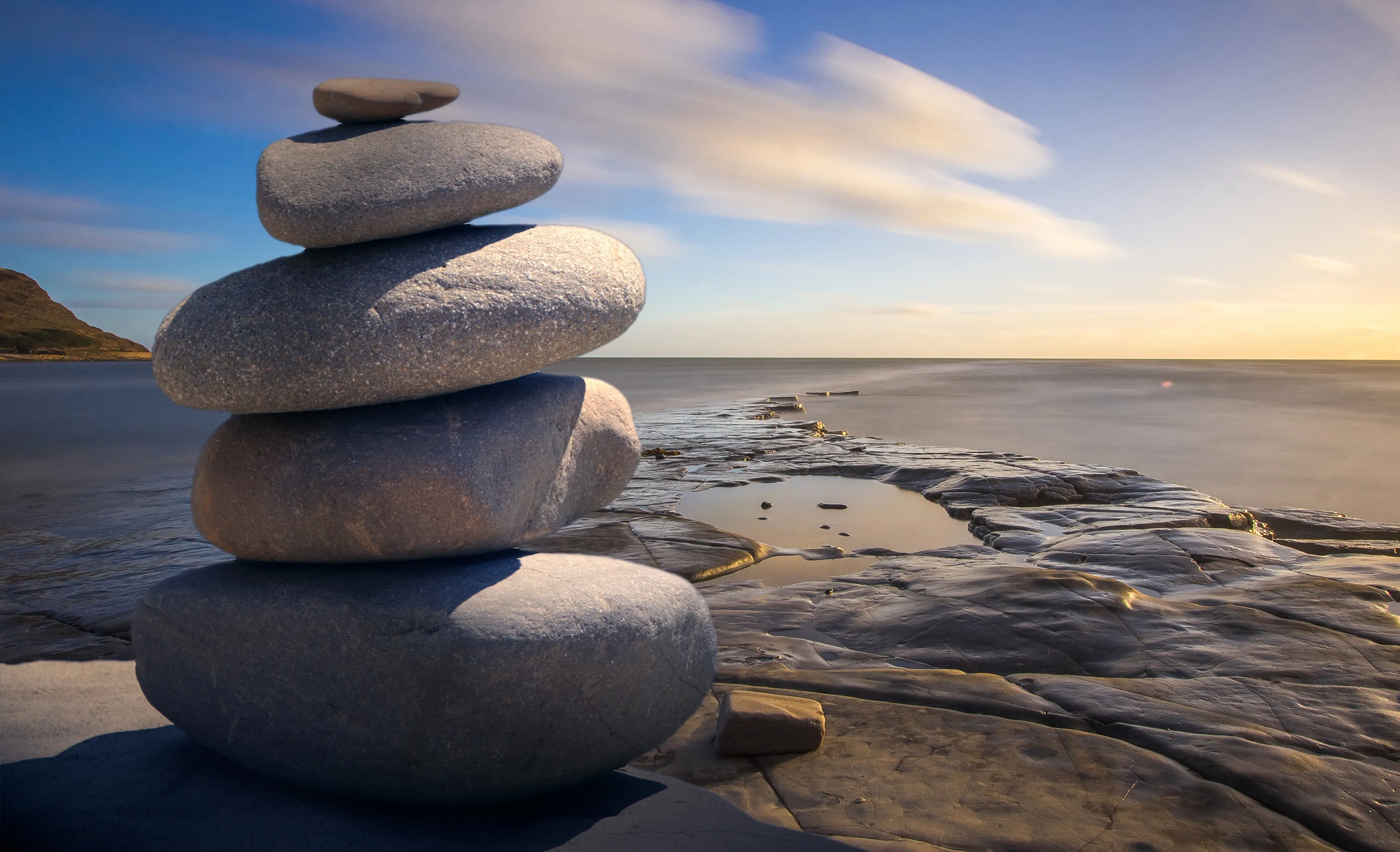 A stack of balanced smooth stones on a rocky shoreline with the sea and a soft sunset sky in the background; blue and earthy tones